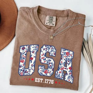 May include: A brown t-shirt with the word "USA" in a floral design. The letters are outlined in blue and filled with a red, white, and blue floral pattern. Below the word "USA" is the text "EST. 1776" in white.