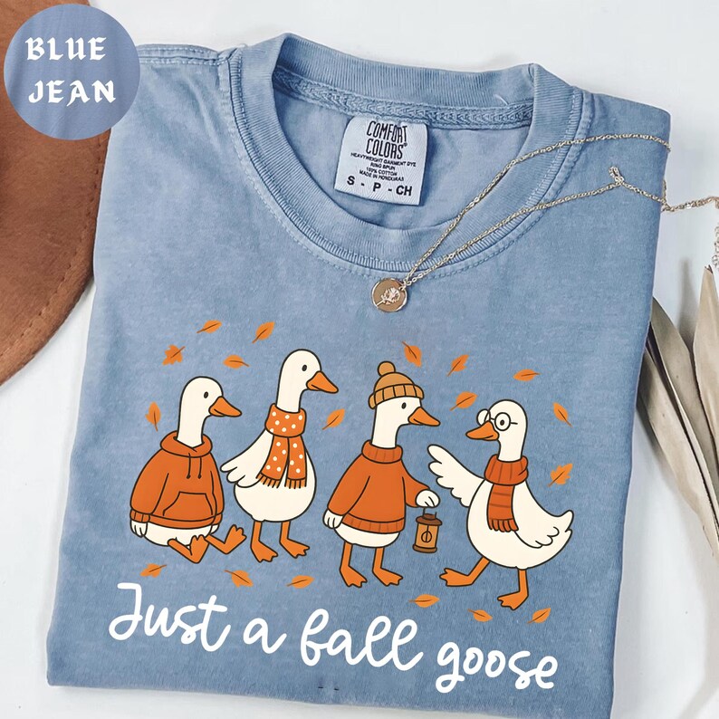Puede incluir: Camiseta azul vaquero con un gr&aacute;fico de cuatro gansos de dibujos animados con atuendos de oto&ntilde;o. Los gansos llevan su&eacute;teres, bufandas y un gorro de color naranja. El texto "Just a fall goose" est&aacute; impreso en la parte inferior.
