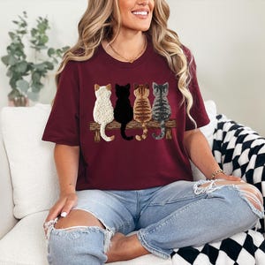 May include: A burgundy t-shirt with a design of four cats sitting on a wooden bench. The cats are white, black, brown, and grey, each with unique patterns. The shirt is a casual, crew neck style.