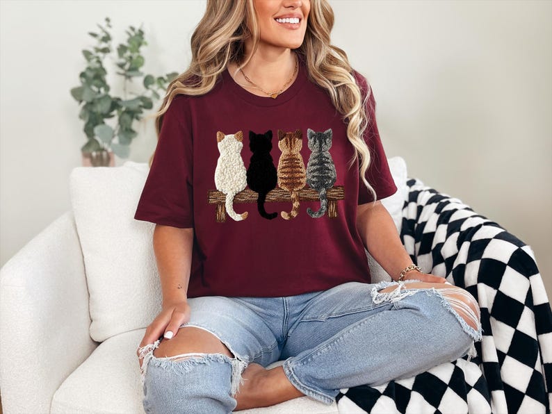May include: A burgundy t-shirt with a design of four cats sitting on a wooden bench. The cats are white, black, brown, and grey, each with unique patterns. The shirt is a casual, crew neck style.