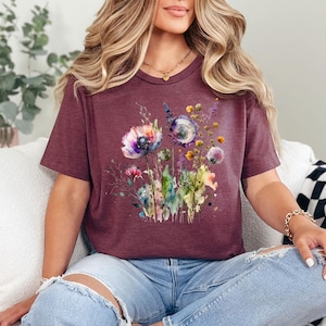 May include: Burgundy t-shirt featuring a watercolor floral design with purple, pink, and yellow flowers. The short-sleeved tee has a crew neck and is made of a soft material. The floral graphic includes various wildflowers and greenery.