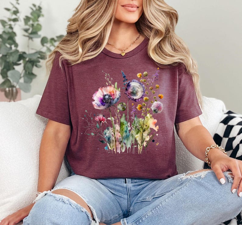 May include: Burgundy t-shirt featuring a watercolor floral design with purple, pink, and yellow flowers. The short-sleeved tee has a crew neck and is made of a soft material. The floral graphic includes various wildflowers and greenery.