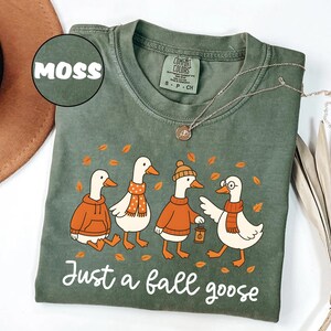 Puede incluir: Camiseta Comfort Colors verde musgo con un gr&aacute;fico de cuatro gansos de dibujos animados con atuendos de oto&ntilde;o. Los gansos llevan su&eacute;teres, bufandas y un gorro. El texto "Just a fall goose" est&aacute; impreso debajo.