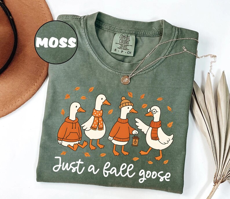 Puede incluir: Camiseta Comfort Colors verde musgo con un gr&aacute;fico de cuatro gansos de dibujos animados con atuendos de oto&ntilde;o. Los gansos llevan su&eacute;teres, bufandas y un gorro. El texto "Just a fall goose" est&aacute; impreso debajo.