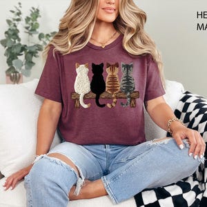 May include: Heather maroon t-shirt with an embroidered design of four cats on a wooden bench. The cats are white, black, brown, and grey. The shirt is a crew neck and is being worn by a person.