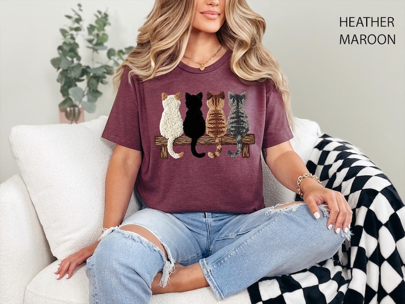 May include: Heather maroon t-shirt with an embroidered design of four cats on a wooden bench. The cats are white, black, brown, and grey. The shirt is a crew neck and is being worn by a person.