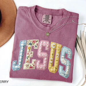 May include: Berry-colored t-shirt with the word "JESUS" in a patchwork design. The letters are in various patterns, including floral, gingham, and polka dots. A silver necklace with a pendant is draped across the shirt.