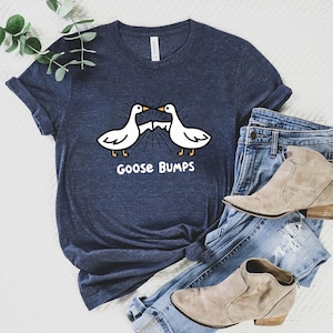 May include: Navy blue t-shirt with a graphic of two white geese facing each other, with the text "Goose Bumps" below. The shirt is paired with blue jeans and tan ankle boots. The shirt is made of a soft material.