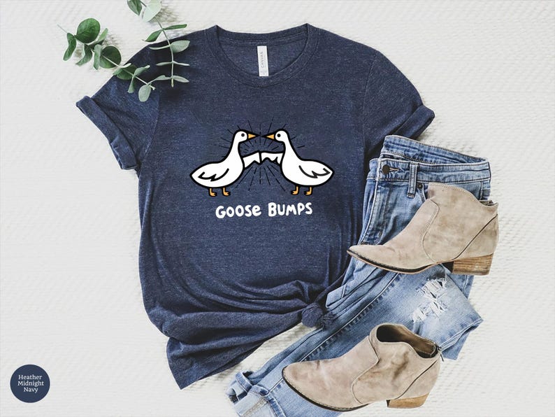 May include: Navy blue t-shirt with a graphic of two white geese facing each other, with the text "Goose Bumps" below. The shirt is paired with blue jeans and tan ankle boots. The shirt is made of a soft material.