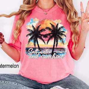 Op de afbeelding: Roze T-shirt met een zonsondergang en palmbomen afbeelding. De tekst "Salt water and sunshine" is op het shirt gedrukt.