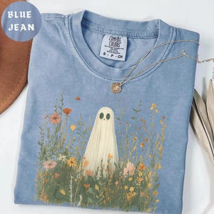 May include: A blue jean colored t-shirt featuring a graphic of a white ghost standing in a field of colorful wildflowers. The shirt has a Comfort Colors brand tag at the neckline and is paired with a thin gold chain necklace with a round pendant.