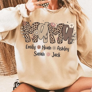 May include: Beige sweatshirt with the word "MAMA" in a mix of leopard print and brown letters. Below the word "MAMA" are the names "Emily", "Noah", "Ashley", "Sarah", and "Jack" with floral accents. The sweatshirt has long sleeves.