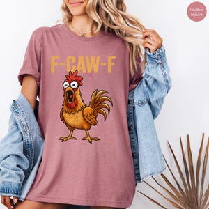 May include: A heather mauve t-shirt featuring a graphic of a wide-eyed, open-mouthed brown chicken with a red comb and wattle. Above the chicken, the text F-CAW-F is printed in a distressed yellow font. The shirt has short sleeves and a crew neck.