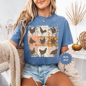 May include: Blue jean t-shirt featuring a patchwork design with brown chicken silhouettes on a floral background. The shirt has short sleeves and a crew neck. The text "BLUE JEAN" is on a blue starburst.