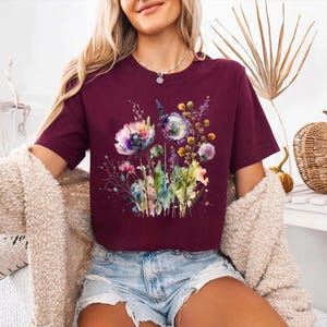 May include: Maroon-colored t-shirt featuring a watercolor floral design with purple, blue, and yellow flowers. The short-sleeved shirt is made of a soft material and is suitable for casual wear. The shirt is paired with denim shorts.