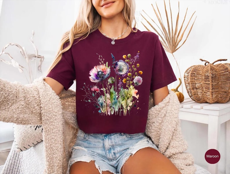 May include: Maroon-colored t-shirt featuring a watercolor floral design with purple, blue, and yellow flowers. The short-sleeved shirt is made of a soft material and is suitable for casual wear. The shirt is paired with denim shorts.