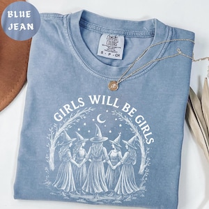 May include: A blue jean Comfort Colors t-shirt with the text "GIRLS WILL BE GIRLS" in white. The graphic features five witch figures holding hands under a crescent moon and stars. A gold necklace with a pendant rests on the shirt.
