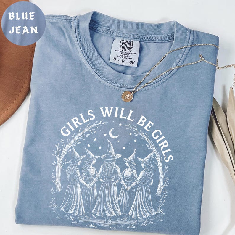 May include: A blue jean Comfort Colors t-shirt with the text "GIRLS WILL BE GIRLS" in white. The graphic features five witch figures holding hands under a crescent moon and stars. A gold necklace with a pendant rests on the shirt.
