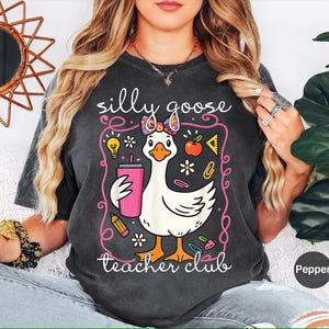 Puede incluir: Camiseta gris oscuro con un dise&ntilde;o caprichoso de un ganso con un lazo y un vaso rosa. El texto dice "silly goose teacher club" en una fuente juguetona, rodeado de ilustraciones escolares.