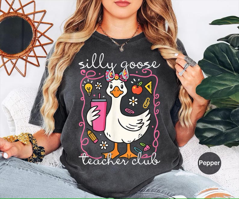 Puede incluir: Camiseta gris oscuro con un dise&ntilde;o caprichoso de un ganso con un lazo y un vaso rosa. El texto dice "silly goose teacher club" en una fuente juguetona, rodeado de ilustraciones escolares.