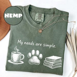 May include: Olive green t-shirt with the text "My needs are simple." and illustrations of a coffee cup, paw print, and books. A gold necklace and the word "HEMP" are also visible. The shirt is a Comfort Colors brand.