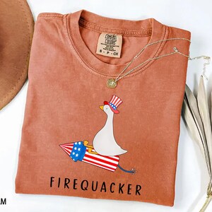 K&ouml;nnte beinhalten: Ein terrakottafarbenes Kurzarm-T-Shirt mit einer Grafik einer wei&szlig;en Gans, die einen rot-wei&szlig;-blau gestreiften Zylinder tr&auml;gt und auf einer Feuerwerksrakete mit Sternen und Streifen reitet. Der Text FIREQUACKER steht unter der Grafik.