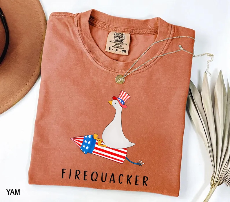 K&ouml;nnte beinhalten: Ein terrakottafarbenes Kurzarm-T-Shirt mit einer Grafik einer wei&szlig;en Gans, die einen rot-wei&szlig;-blau gestreiften Zylinder tr&auml;gt und auf einer Feuerwerksrakete mit Sternen und Streifen reitet. Der Text FIREQUACKER steht unter der Grafik.