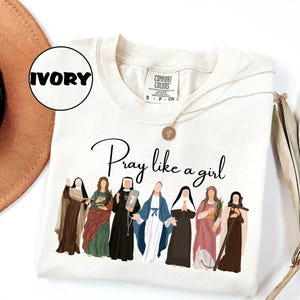 May include: Ivory-colored t-shirt with the phrase "Pray like a girl" in black script. The shirt features an illustration of seven women in religious attire. A gold necklace with a pendant is draped across the shirt.