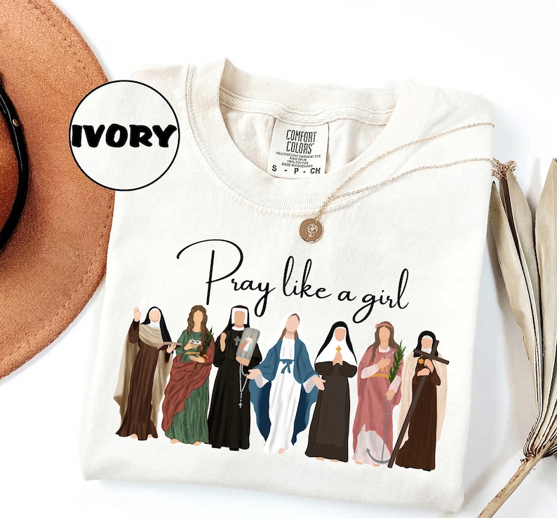 May include: Ivory-colored t-shirt with the phrase "Pray like a girl" in black script. The shirt features an illustration of seven women in religious attire. A gold necklace with a pendant is draped across the shirt.