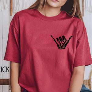 Pode incluir: Uma camiseta vermelha de manga curta com uma m&atilde;o de esqueleto preta fazendo um sinal shaka.