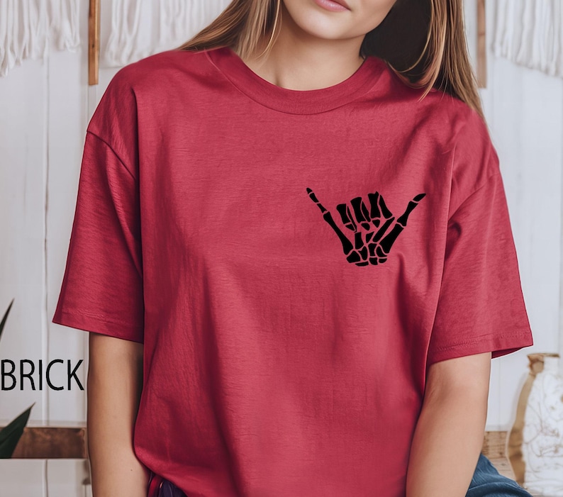 Pode incluir: Uma camiseta vermelha de manga curta com uma m&atilde;o de esqueleto preta fazendo um sinal shaka.