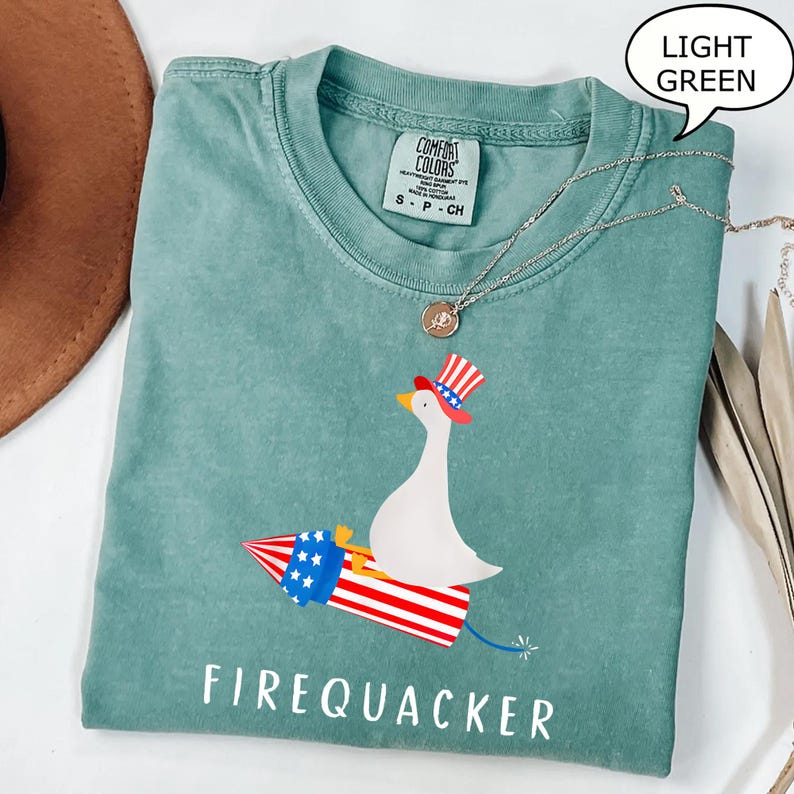 K&ouml;nnte beinhalten: Ein hellgr&uuml;nes T-Shirt mit einer Grafik einer wei&szlig;en Gans, die einen rot-wei&szlig;-blauen Zylinder tr&auml;gt und auf einer Feuerwerksrakete reitet. Der Text FIREQUACKER ist unter der Grafik gedruckt. Das T-Shirt hat ein Comfort Colors Markenetikett.