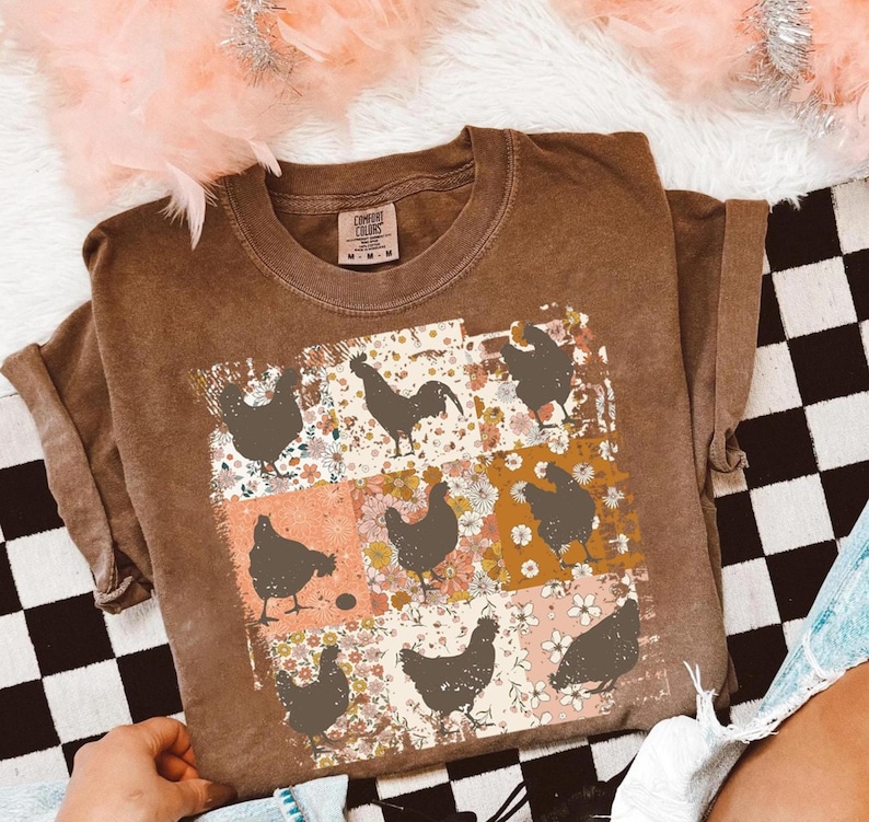May include: Brown short-sleeved t-shirt with a crew neck. The shirt features a patchwork design of floral patterns and chicken silhouettes. The shirt has a Comfort Colors tag.