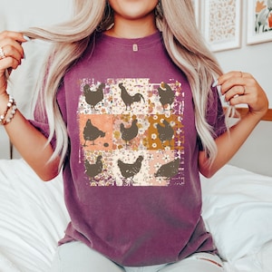 May include: A purple t-shirt featuring a patchwork design of chickens and floral patterns. The design includes various chicken silhouettes in brown against a backdrop of orange, white, and brown floral squares. The shirt has short sleeves.