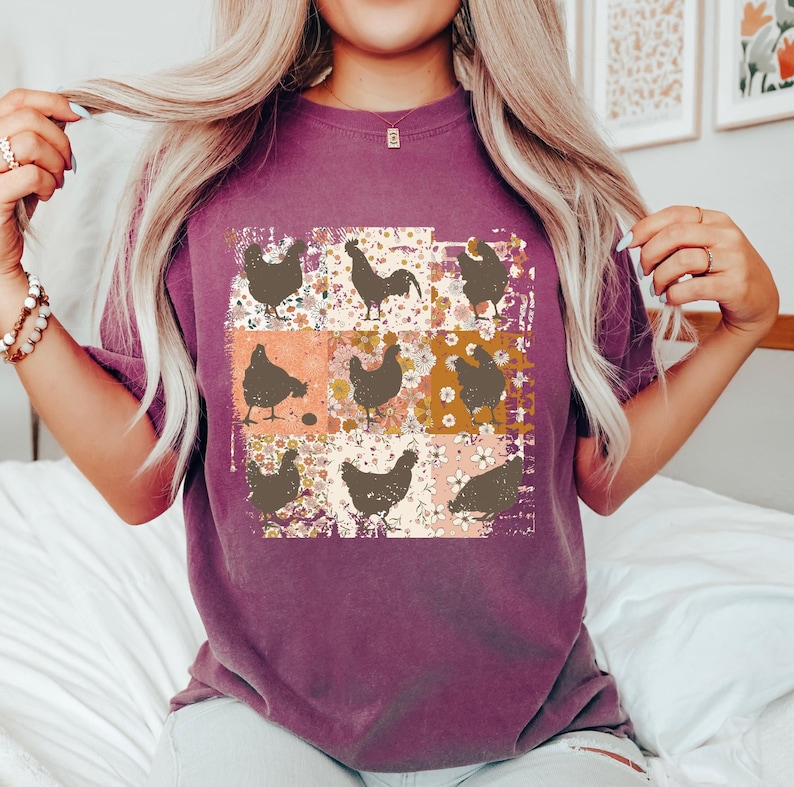 May include: A purple t-shirt featuring a patchwork design of chickens and floral patterns. The design includes various chicken silhouettes in brown against a backdrop of orange, white, and brown floral squares. The shirt has short sleeves.