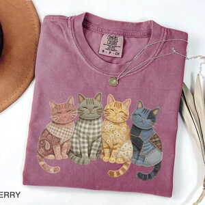 May include: Berry-colored t-shirt featuring four whimsical cat illustrations with patchwork patterns. The cats have different colors and textures, with smiling faces. A silver necklace with a pendant is on the shirt. The shirt has a Comfort Colors tag.