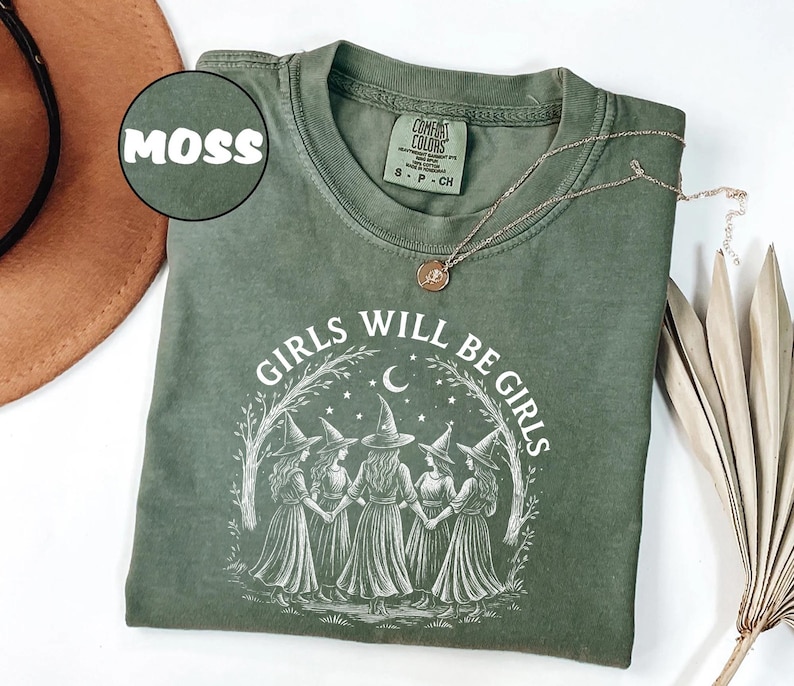 May include: A moss green t-shirt featuring the text "GIRLS WILL BE GIRLS" and an illustration of five witches. The shirt has a crew neck and short sleeves. A gold necklace with a pendant is draped across the shirt.