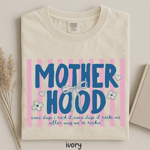 May include: Ivory-colored t-shirt with the word "MOTHERHOOD" in blue, set against pink vertical stripes. The design includes a bow, hearts, and flowers. The text "some days i rock it, some days it rocks me either way we're rockin'" is also included.