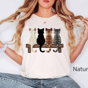 May include: Cream-coloured t-shirt with an embroidered design of four cats on a wooden bench. The cats are white, black, brown, and grey. The word "Natural" is printed in black on the lower right.