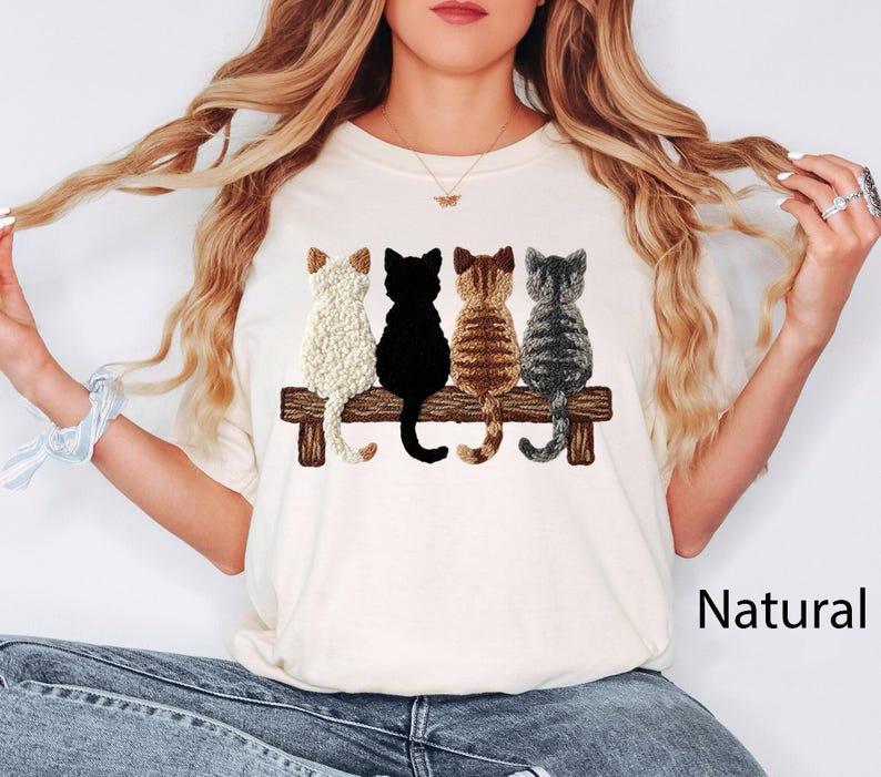 May include: Cream-coloured t-shirt with an embroidered design of four cats on a wooden bench. The cats are white, black, brown, and grey. The word "Natural" is printed in black on the lower right.