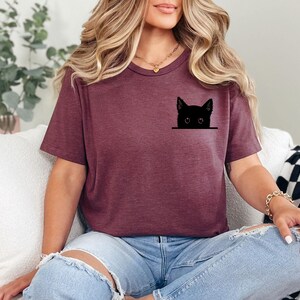 May include: A maroon short-sleeved t-shirt featuring a black graphic of a cat head peeking out from the chest area. The cat design is simple and minimalist, showing only the ears, eyes, and top of the head in a solid black silhouette style.