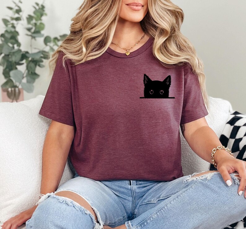 May include: A maroon short-sleeved t-shirt featuring a black graphic of a cat head peeking out from the chest area. The cat design is simple and minimalist, showing only the ears, eyes, and top of the head in a solid black silhouette style.