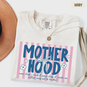 May include: Ivory-colored t-shirt with the word "MOTHERHOOD" in blue, set against pink vertical stripes. The shirt includes the text "some days i rock it, some days it rocks me either way we're rockin'". A gold necklace is draped across the shirt.