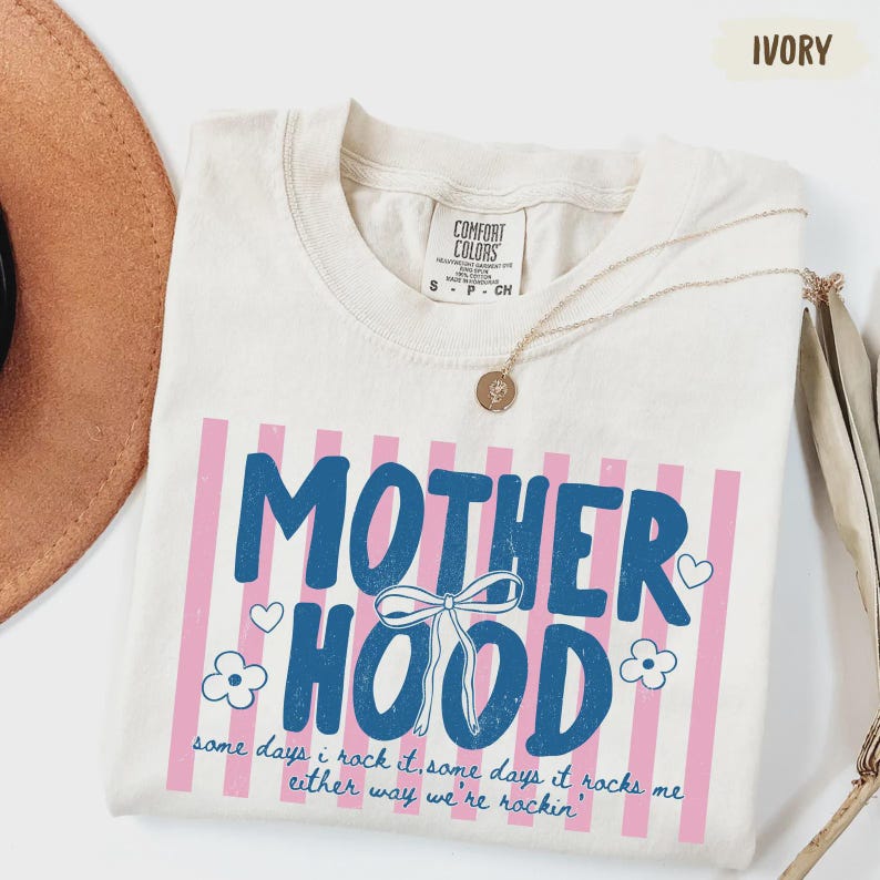 May include: Ivory-colored t-shirt with the word "MOTHERHOOD" in blue, set against pink vertical stripes. The shirt includes the text "some days i rock it, some days it rocks me either way we're rockin'". A gold necklace is draped across the shirt.