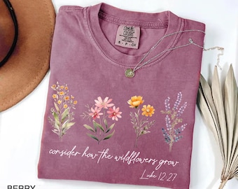 May include: Berry-colored t-shirt with a floral design and the text "consider how the wildflowers grow" and "Luke 12:27". The shirt has a crew neck and short sleeves. A silver necklace with a pendant is on the shirt.