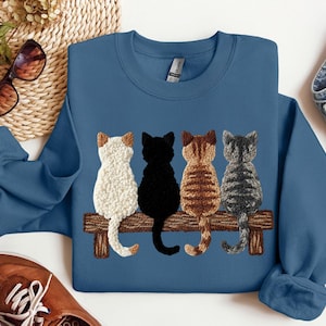 May include: Indigo blue sweatshirt with an embroidered design of four cats on a wooden bench. The cats are white, black, brown, and grey. The sweatshirt has a crew neck and long sleeves.