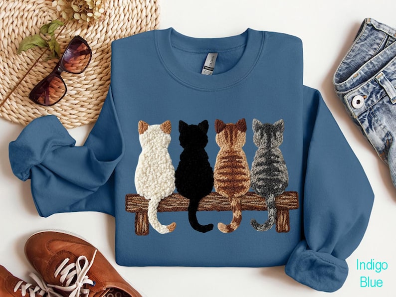 May include: Indigo blue sweatshirt with an embroidered design of four cats on a wooden bench. The cats are white, black, brown, and grey. The sweatshirt has a crew neck and long sleeves.