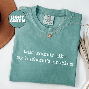 May include: Light green Comfort Colors t-shirt with the phrase "that sounds like my husband's problem" in white. A silver necklace with a small pendant rests on the shirt. The shirt is folded on a white surface.