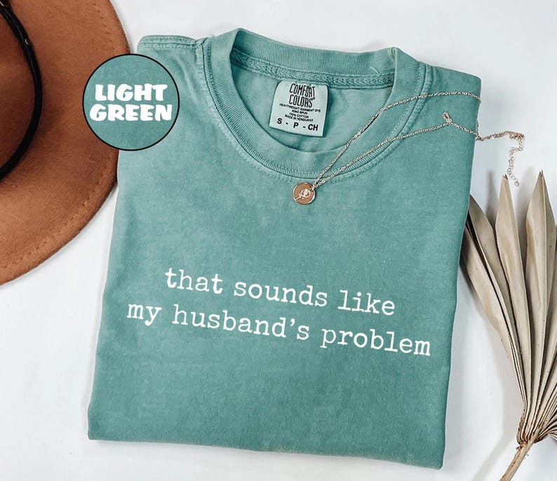 May include: Light green Comfort Colors t-shirt with the phrase "that sounds like my husband's problem" in white. A silver necklace with a small pendant rests on the shirt. The shirt is folded on a white surface.