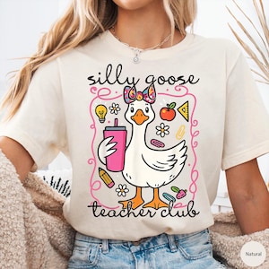 Puede incluir: Camiseta color crema con un dise&ntilde;o de ganso de dibujos animados con un lazo y sosteniendo una bebida rosa. El texto dice "silly goose teacher club". El dise&ntilde;o incluye elementos escolares como una bombilla, una manzana y un l&aacute;piz.